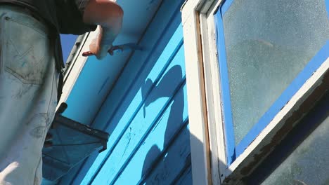 Do-it-yourself-house-painting-in-Waco,-Texas-in-USA,-steady-shot-from-underneath-of-man-painting-blue-colour-on-wooden-house-in-slow-motion