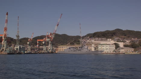 Sailing-past-port-cranes-and-a-warship-that-is-docked-at-Kobe-Port,-Japan