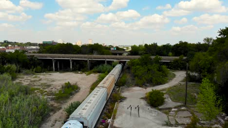 A-drone-shot,-with-downward-pedestal-motion,-capturing-horizontal-highways-in-distance-and-a-goods-train-passing-from-under-them-and-trees-sorrounding-the-entire-scene-and-a-bright-blue-sky