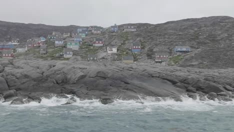 Sailing-Past-Rocky-Outcrop-With-Waves-Crashing-And-Buildings-In-Perched-On-Hill-In-The-Background-In-Greenland