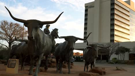 Panning-shot-of-three-cattle-statues-in-a-park-with-cars-driving-across-and-a-building-in-the-background,-shot-on-a-sunny-day