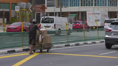 Hombre-Singapurense-Empujando-Un-Carrito-De-Cajas-De-Cartón-En-Medio-De-La-Avenida---Plano-De-Seguimiento-Amplio