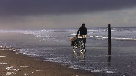 Old-fisherman-walking-his-bike-down-the-mudflat-shoreline-in-Texel-Island-Beach,-North-Holland,-the-Netherlands---Wide-Static-shot