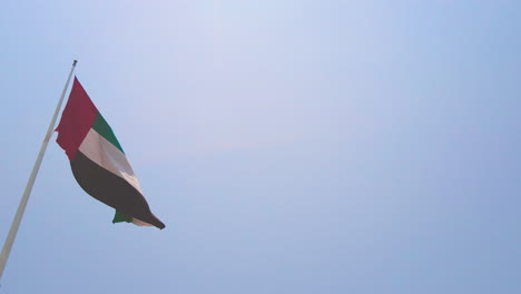 Big-UAE-flag-on-pole-in-Abu-Dhabi-waving-slowly-in-the-wind-on-bright-sunny-day-with-clear-blue-sky-close-bottom-up-land-view
