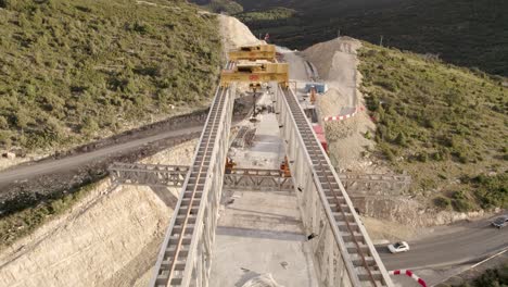Backward-fly-by-motion-video-over-a-new-bridge´s-structure-under-construction-in-Barranco-de-la-Bota-in-Morella,-showing-cranes-and-railways-from-above