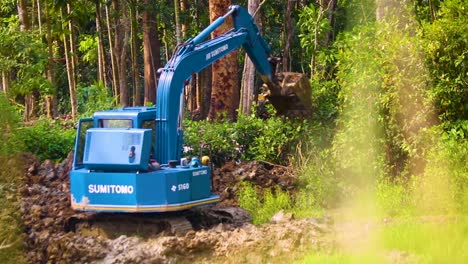 Blue-Excavator-Dropping-Soil-and-Rotating-at-the-Edge-of-a-Rainforest-being-Deforested,-Shot-through-High-Grass-Shoots-Moving-in-the-Wind