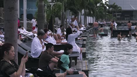 Tourist-guests-resting-on-Lounge-Chairs-by-the-edge-of-infinity-Pool-at-Marina-Bay-Sands-Hotel-SkyPark,-Singapore---Wide-static-side-view