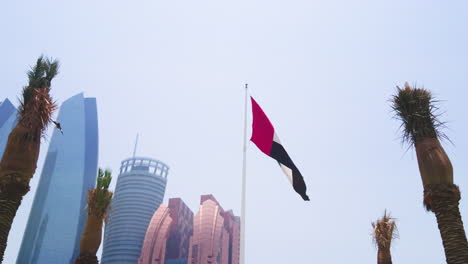 Big-UAE-flag-waving-slowly-in-the-wind-and-falcon-flying-in-front-of-it-framed-centered-by-palm-trees-with-Bab-Al-Qasr-Hotel-and-Etihad-Towers-in-backgroung-on-Abu-Dhabi-Corniche-Street-close-day-view