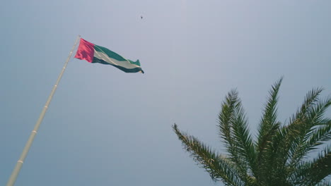 Große-VAE-Flagge-An-Der-Stange-In-Abu-Dhabi,-Die-An-Einem-Strahlend-Sonnigen-Tag-Langsam-Im-Wind-Weht,-Mit-Klarem,-Blauem-Himmel-Und-Blick-Von-Unten-Nach-Oben-Auf-Das-Land