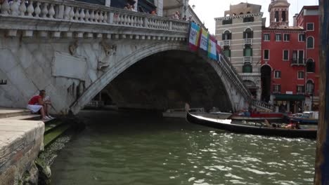 Venetian-gondola-with-tourist-riding-under-the-Rialto-Bridge-in-a-sunny-day-in-Venice,-Italy