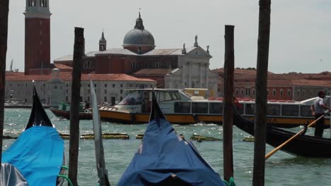 Venetian-gondolas-docking-in-a-pier-with-boats-passing-by-and-the-bell-tower-of-St