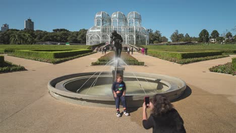 Tourists-visiting-the-Botanical-Garden-of-Curitiba,-in-the-foreground-the-statue-of-"Maternal-Love"-and-in-the-background-the-"Crystal-Palace",-timelapse-capture