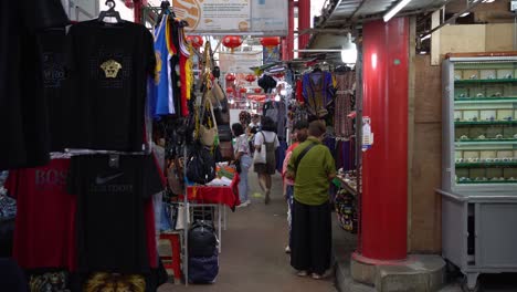 Point-of-view-of-a-lady-walking-through-a-narrow-pathway-of-stalls-and-other-shoppers-shopping-at-a-famous-landmark-marketplace,-Petaling-Street,-Kuala-Lumpur,-Malaysia