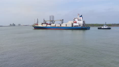 Ein-Riesiges-Containerschiff-Beherrscht-Die-Bucht,-Während-Es-Sich-Elegant-Einem-Industriehafen-Nähert,-Gefolgt-Von-Einem-Speziellen-Schlepper,-Der-Dicht-Dahinter-Folgt,-Mit-Malerischer-Aussicht,-Industriehafen,-Logistik,-Containerschiff