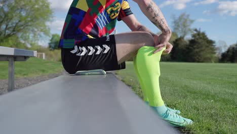 Cinematic-slowmotion-Soccer-player-with-tattoos-sitting-on-bench-getting-ready-to-play-game-putting-on-shin-guards-with-colorful-socks-spring-green-soccer-field-team-player-practicing-dribble-kick