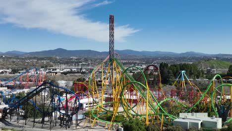 Aerial-left-to-right-shot-of-guests-riding-many-colorful-rollercoasters-at-Six-Flags-Magic-Mountain-theme-park-located-in-Santa-Clarita,-California-on-a-summer-day-with-city-and-hills-in-background
