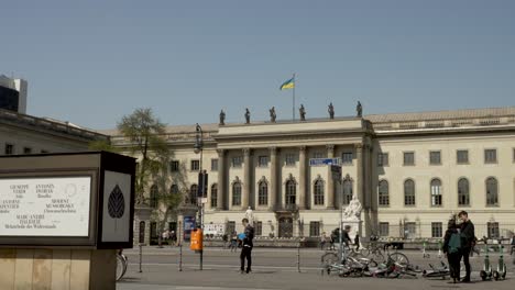 Traffic-And-People-Going-Past-View-Of-Humboldt-University-of-Berlin-On-Sunny-Day-With-Blue-Skies-And-Ukraine-Flag-Fluttering-On-Top