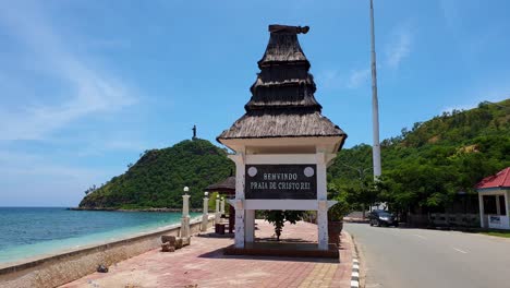 Welcome-to-Cristo-Rei-beach-sign-with-traditional-Timorese-house-and-Cristo-Rei-statue-in-capital-Dili,-Timor-Leste