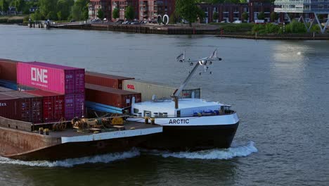 Aerial-Close-Up-View-Off-Forward-Bow-Of-Arctic-Cargo-Container-Ship-Paired-With-Barge-Travelling-Along-River-Noord
