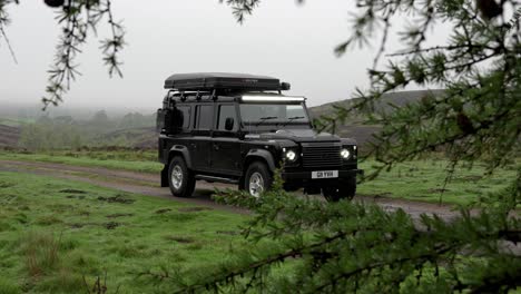 Black-modified-camping-land-rover-defender-with-rooftop-tent-parked-static-on-a-single-lane-country-road-on-cloudy-moody-day-using-trees-and-green-as-foreground