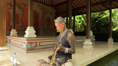 Slow-motion-parallax-shot-of-a-traditional-snake-charmer-in-a-balinese-temple-wearing-a-snake-around-his-neck-and-elegantly-controlling-it-in-bali-indonesia