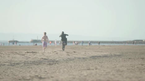 Adult-on-dirt-bike-riding-on-sandy-beach-doing-wheelies-with-people-near
