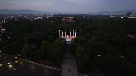 Aerial-view-in-front-of-the-illuminated-Altar-a-la-Patria-with-Castillo-de-Chapultepec-in-the-background,-dusk-in-CDMX
