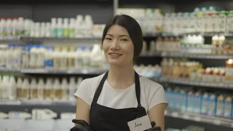 Porträt-Einer-Attraktiven-Jungen-Asiatischen-Verkäuferin-In-Schwarzer-Schürze,-Die-In-Einem-Supermarkt-Mit-Regalen-Voller-Milchprodukte-Steht