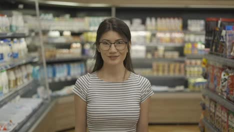 Retrato-De-Una-Joven-Y-Atractiva-Mujer-Asiática-Parada-En-Un-Supermercado-Con-Estantes-De-Productos-Lácteos-Al-Fondo,-Mirando-La-Cámara-Y