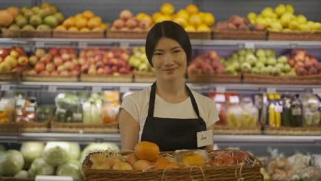 Retrato-De-Una-Atractiva-Joven-Trabajadora-Asiática-Parada-En-Un-Supermercado-Con-Estantes-De-Frutas-En-El-Fondo,-Mirando-La-Cámara-Y-Sonriendo.-Concepto-De-Negocio-Y-Personas-De-Comercio