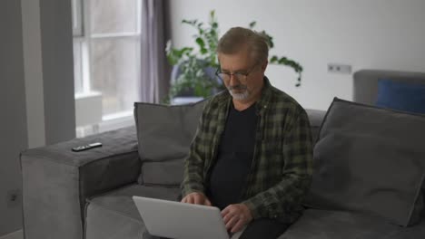 Älterer-älterer-Reifer-Mann,-Der-Auf-Dem-Laptop-Tippt-Und-Im-Internet-Surft,-Sitzt-Allein-Auf-Dem-Sofa