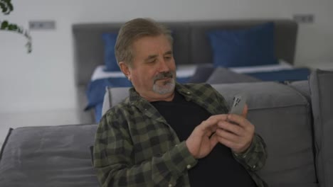 Smiling-senior-grey-haired-man-sitting-on-couch-and-using-mobile-phone-at-home
