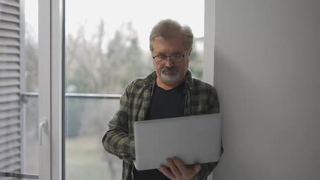 Senior-older-mature-man-typing-on-laptop-browsing-internet-standing-next-to-the-window