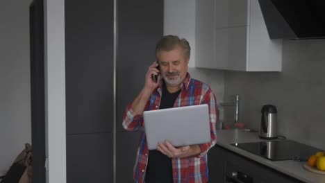 Bearded-old-man-using-laptop-in-kitchen-and-talking-by-phone-at-the-same-time
