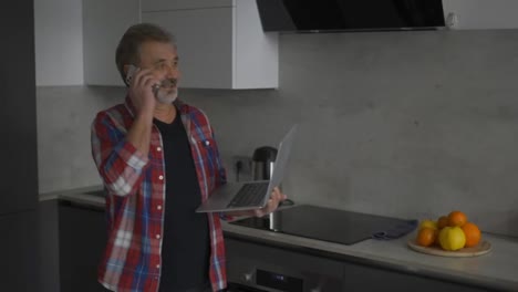Moderner-Mann-Im-Alter-Benutzt-Laptop-In-Der-Küche-Und-Telefoniert-Gleichzeitig