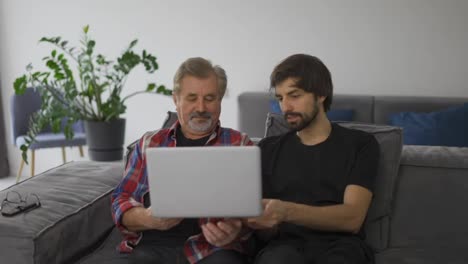 Young-man-teaching-old-dad-using-internet-online-with-computer-on-couch-in-living-room