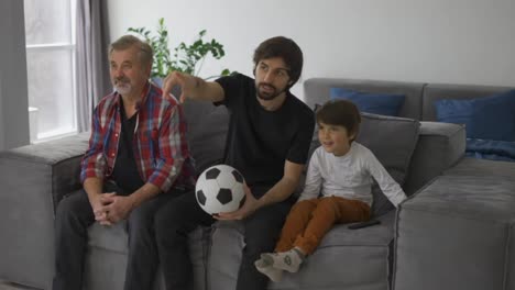 Three-generations-of-men-relax-on-couch-in-living-room-watch-football-match