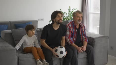 Three-generations-of-men-on-couch-in-living-room-watch-football-match