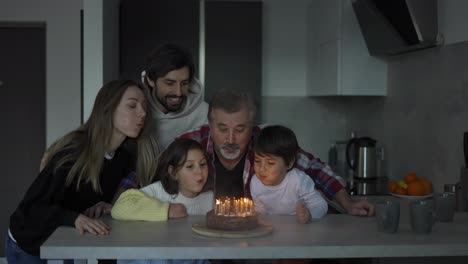 Glücklicher-Großvater-Bläst-Kerzen-Auf-Geburtstagstorte,-Umgeben-Von-Seiner-Familie