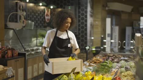 Multiracial-worker-in-black-apron-stocking-yellow-peppers-in-supermarket