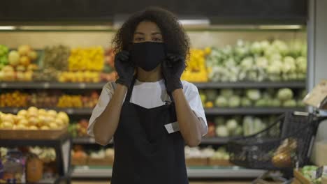 Portrait-of-happy-African-American-female-worker-in-black-mask-and-gloves-standing-in-supermarket,-taking-off-protection-mask