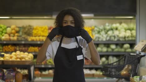 Portrait-of-happy-African-American-female-worker-standing-in-supermarket,-putting-on-protection-mask