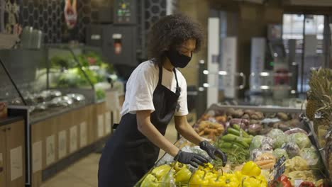 Mitarbeiter-Eines-Gemischtrassen-Lebensmittelladens-Mit-Gesichtsmaske-Und-Handschuhen,-Der-Frisches-Gemüse-Und-Obst-Hineinlegt