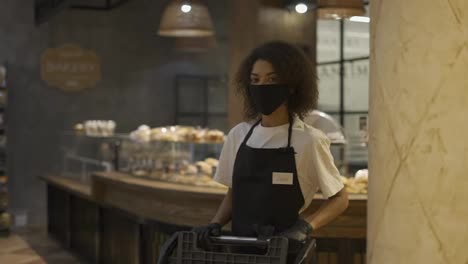 African-American-female-worker-pushing-trolley-in-supermarket,-wearing-protection-mask