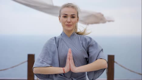 Portrait-of-smiling-female-coach-yoga-in-robe-standing-at-terrace-folded-hands-in-Namaste-gesture