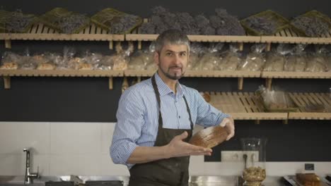 Handsome-man-in-apron-taking-fresh-bread-from-the-shelf-and-showing-it-on-camera.-Indoor,-bakery-store