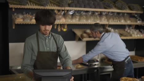 Young-cashier-in-apron-typing-on-touch-screen,-handsome-bakery-worker-slicing-bread-in-the-background