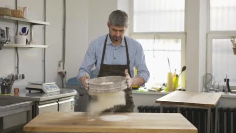 Handsome-baker-sifting-flour-through-a-sieve-at-the-kitchen.-Close-up,-indoor,-slow-motion