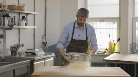 Handsome-male-baker-sifting-flour-through-a-sieve-at-the-kitchen.-Close-up,-indoor,-slow-motion