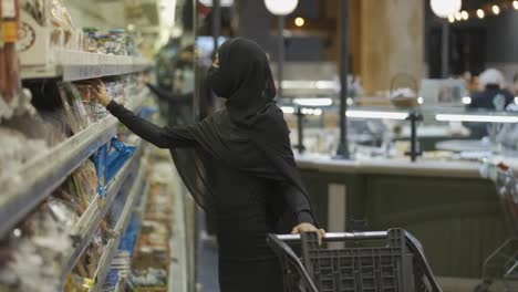 Woman-in-hijab-and-protective-mask-doing-shopping,-takes-product-from-the-shelf
